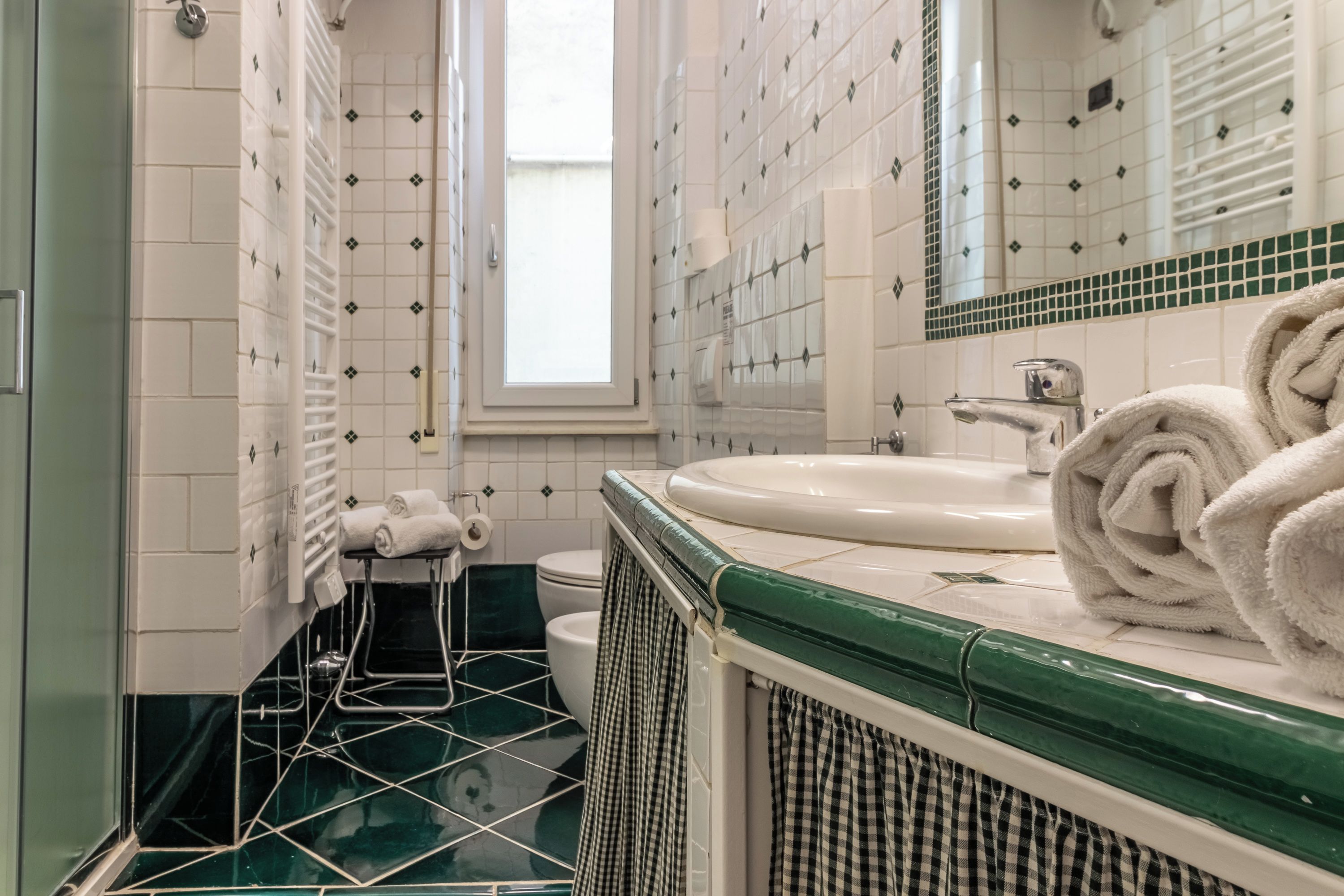 Un bagno con un lavandino bianco, pavimento piastrellato in verde e uno specchio sopra il lavandino.