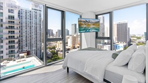 A bedroom with a large bed, a TV mounted on the wall, and a city view through large windows. - 2-bedroom condo in marvelous Miami Views in the perfect spot! (Miami)