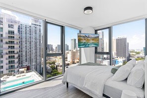 Ein Schlafzimmer mit einem großen Bett, einem an der Wand montierten Fernseher und Blick auf die Stadt durch große Fenster.
