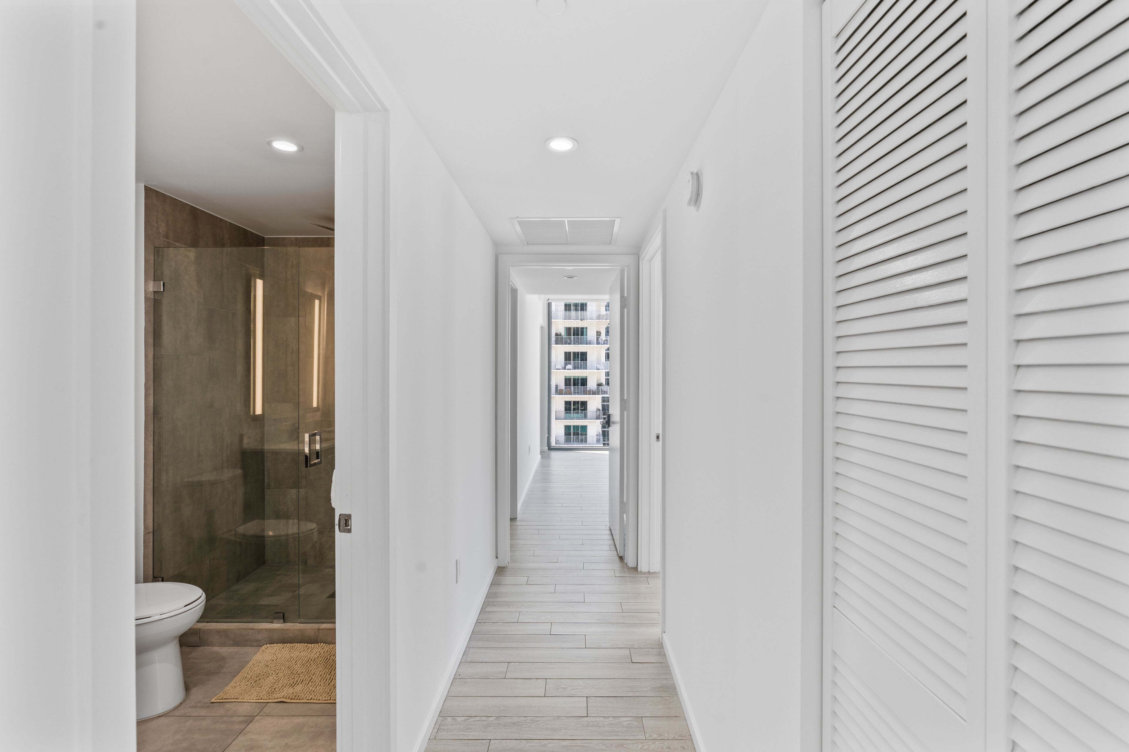 A narrow hallway with a glass-enclosed shower, a white toilet, and a wooden floor.