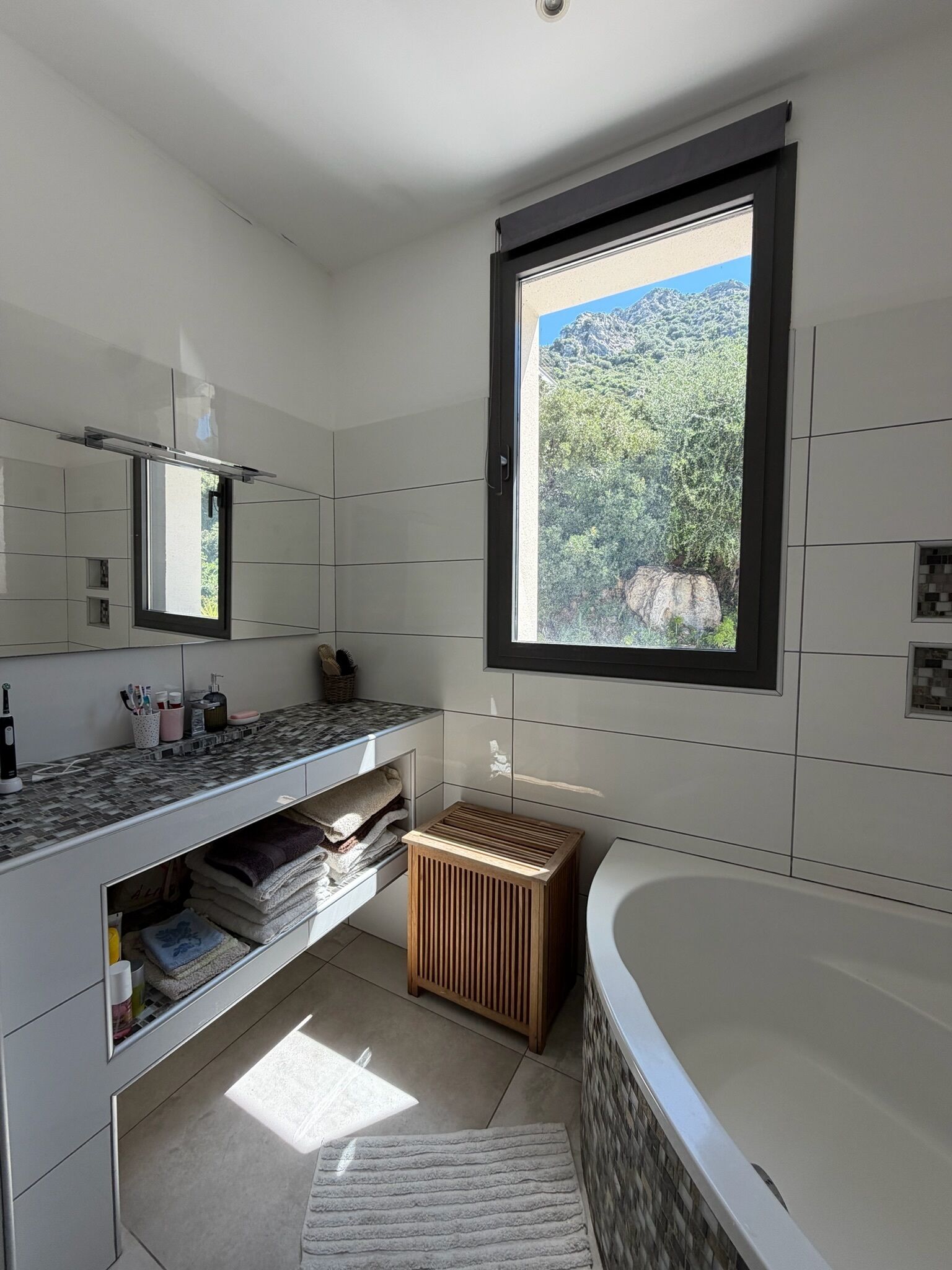 Une salle de bain moderne dotée d’une grande baignoire, d’un meuble-lavabo avec miroir et d’une fenêtre offrant une vue sur un paysage montagneux.
