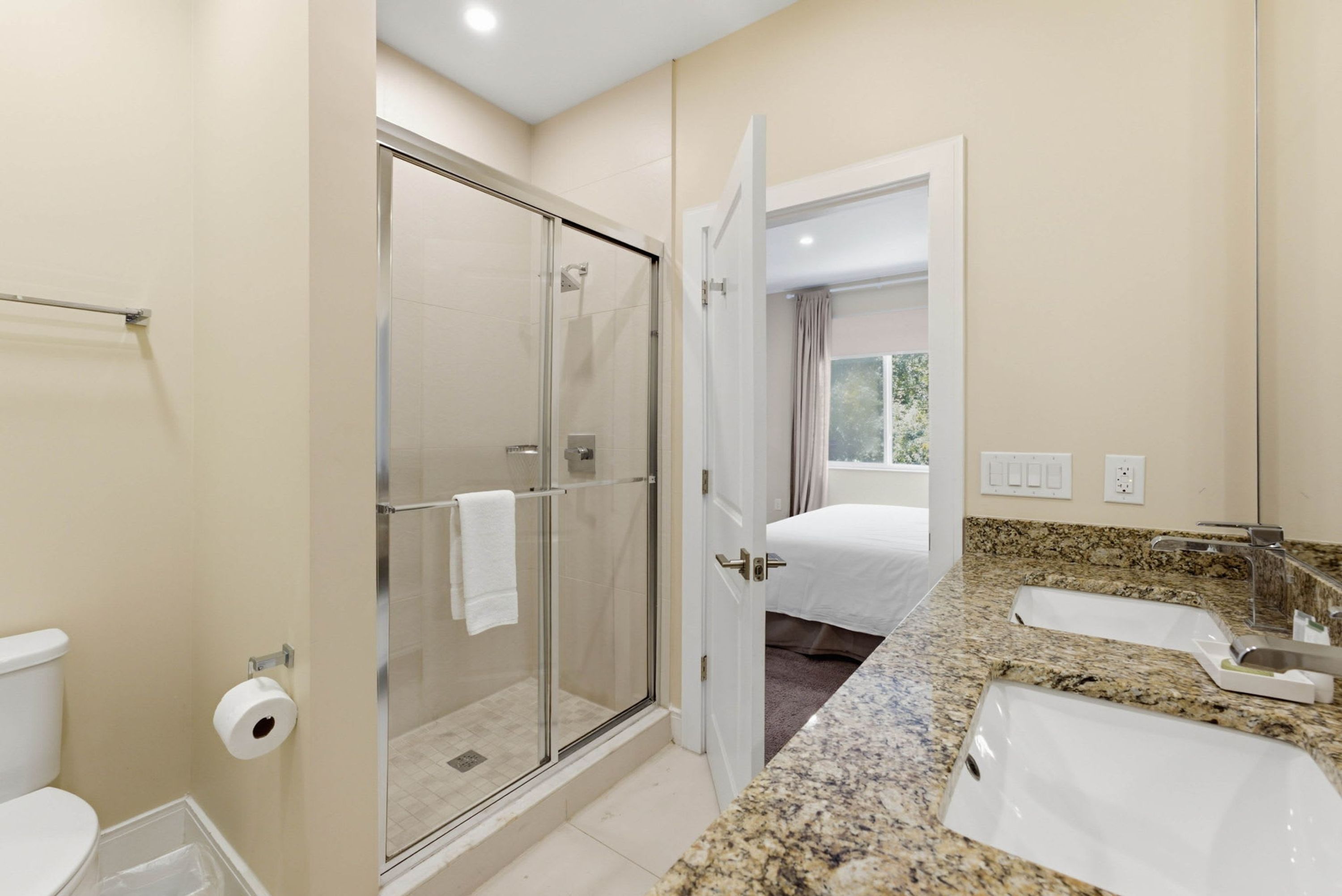 A modern bathroom with a glass-enclosed shower, a toilet, a sink, and a bed visible through an open door.