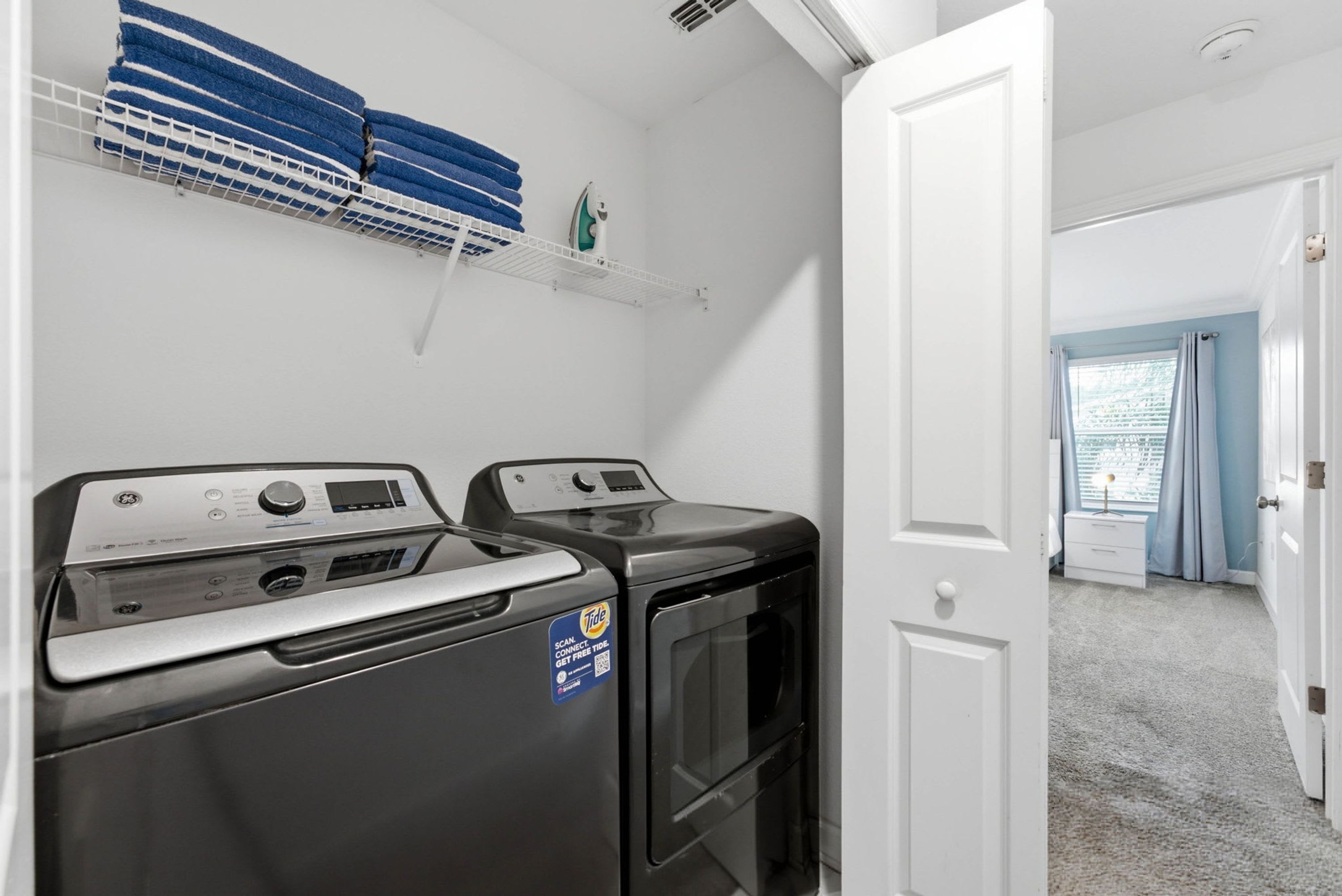 A laundry room with two front-loading washing machines, a shelf with folded towels, and a door leading to another room.