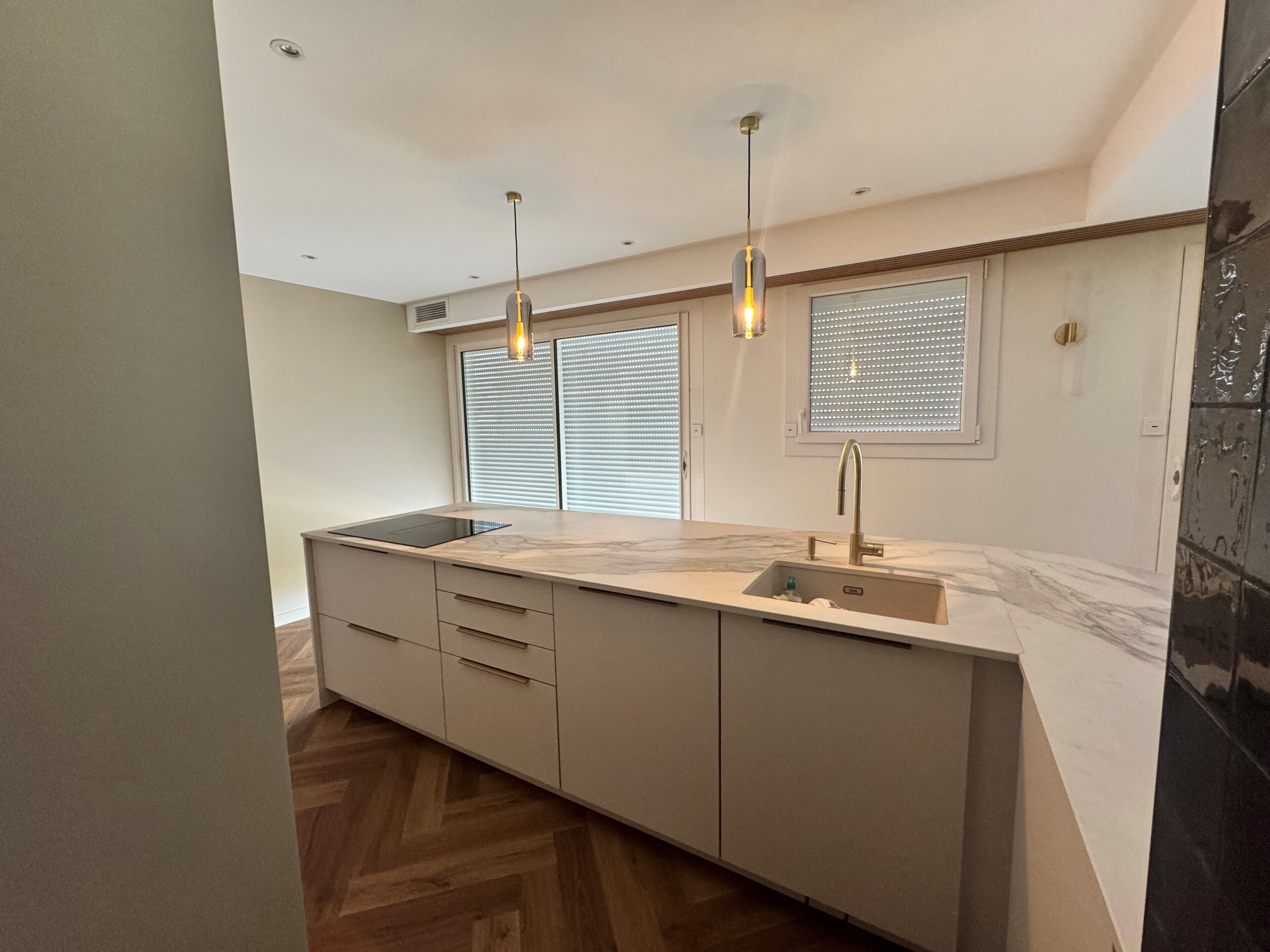 Een moderne keuken met een centraal eiland, hangende verlichting en een raam met jaloezieën.