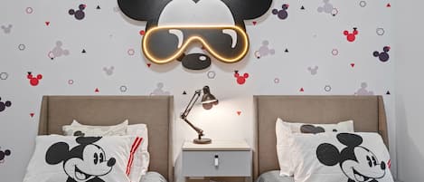 Un dormitorio con dos camas, cada una con almohadillas de Mickey Mouse, una mesita de noche con lámpara y un adorno de pared con la cabeza de Mickey Mouse.