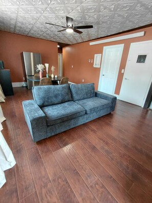 A room with a grey sofa, wooden flooring, a ceiling fan, and a white door.