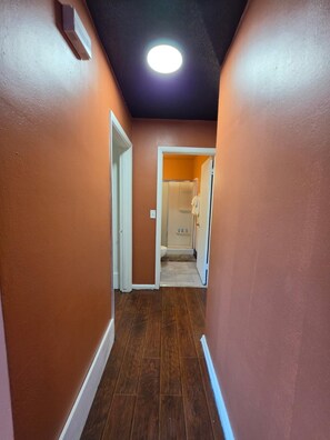 A hallway with wooden flooring and orange walls, leading to a bathroom with a shower and toilet.