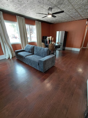 A spacious room with a grey sofa, wooden flooring, a ceiling fan, and a window with curtains.