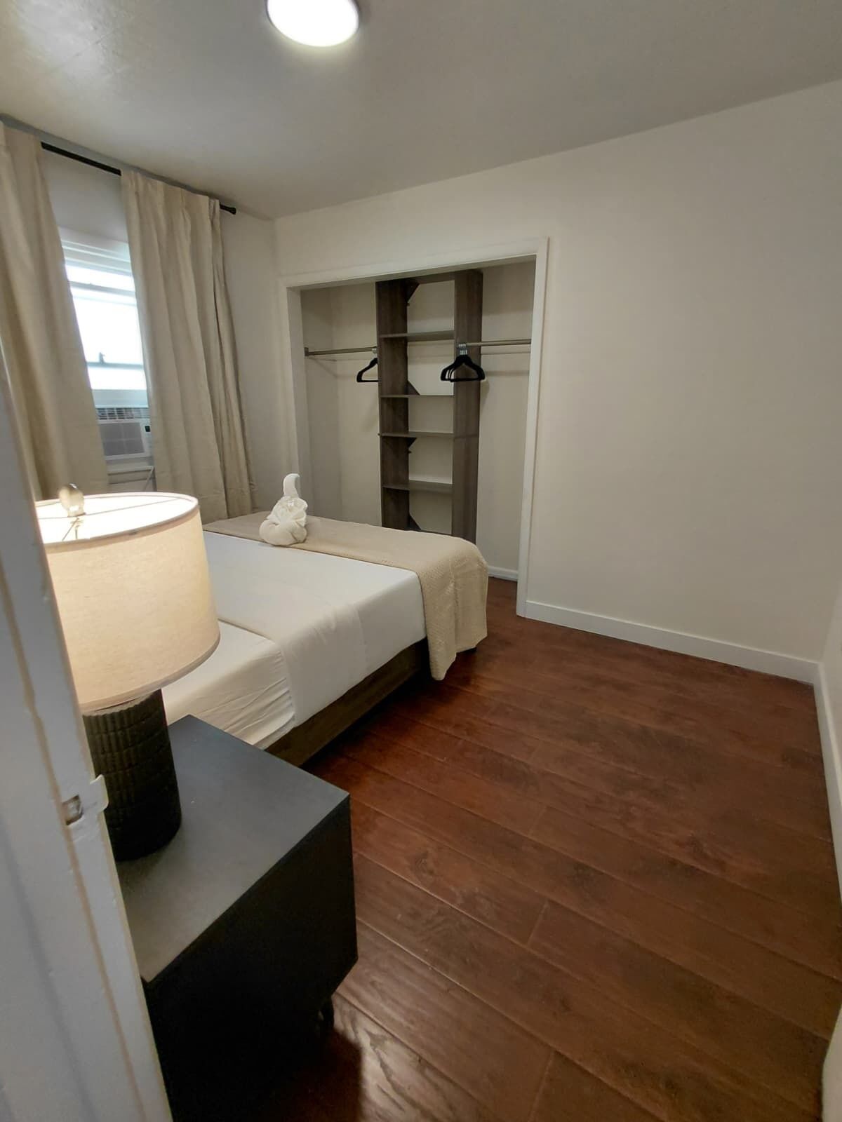 Ein Schlafzimmer mit Bett, Nachttisch mit Lampe, Kleiderschrank und Blick durch ein Fenster.