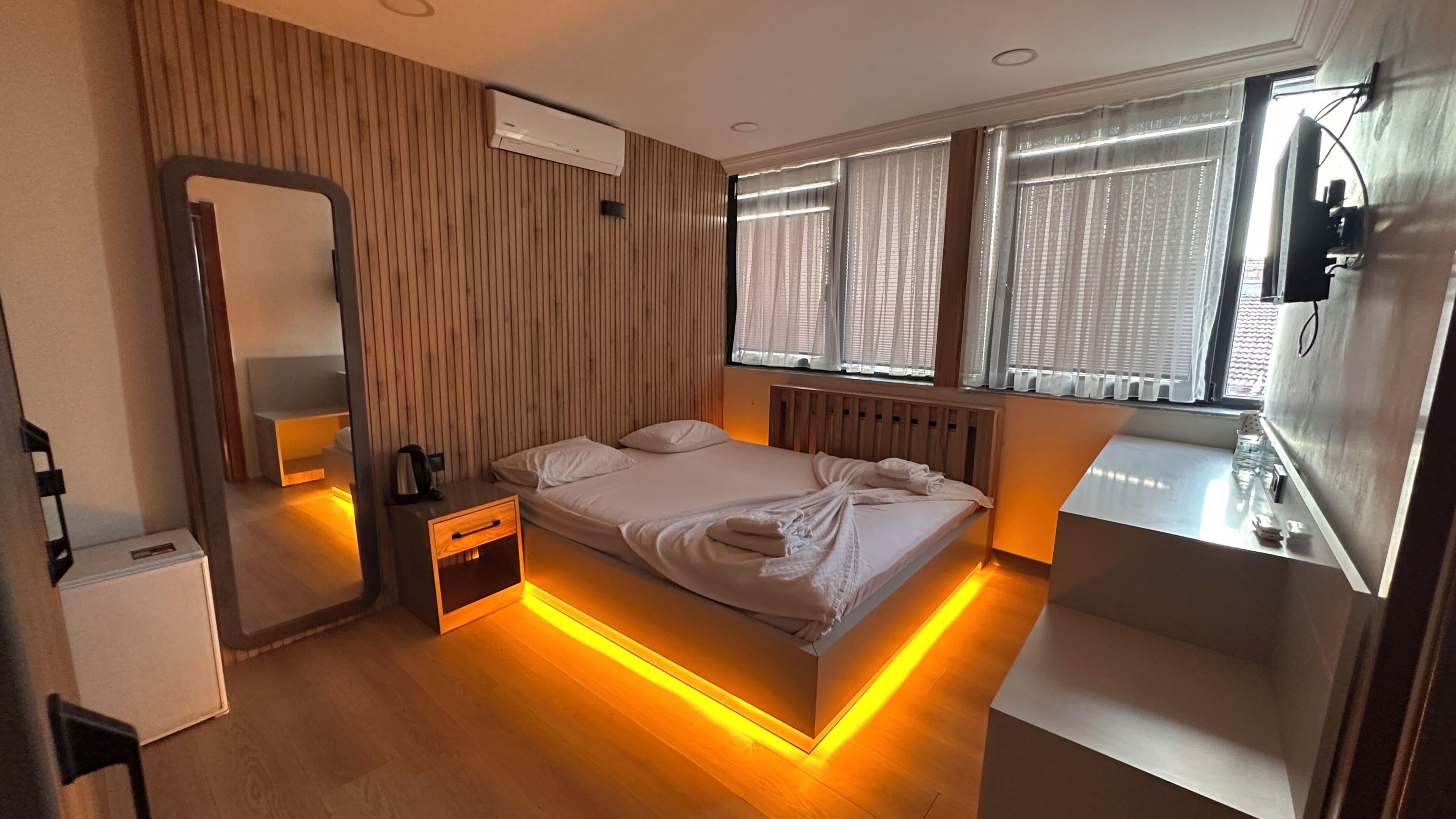 Ett modernt hotellrum med en sänggavel i trä, ett nattduksbord med en vattenkokare, en stor spegel och en nattlampa.