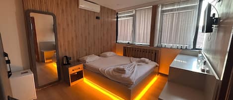 A modern hotel room with a wooden headboard, a bedside table with a kettle, a large mirror, and a bedside lamp.