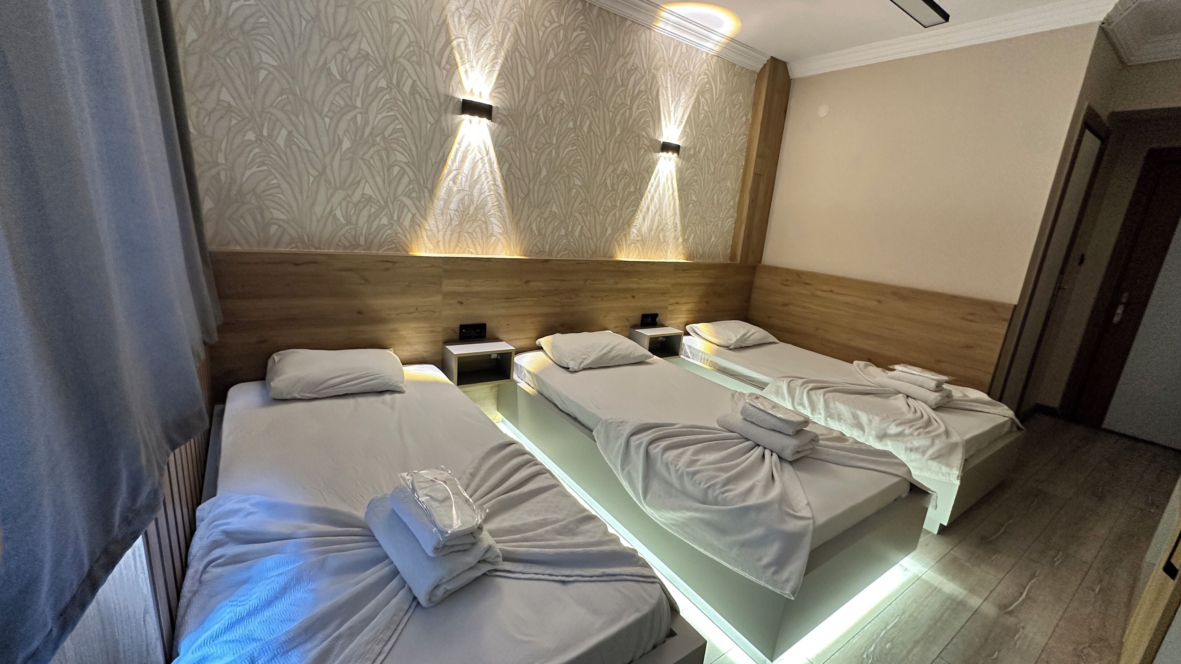 A hotel room with three single beds, each with a neatly folded towel and a pillow. The room has a wooden headboard and a patterned wallpaper.