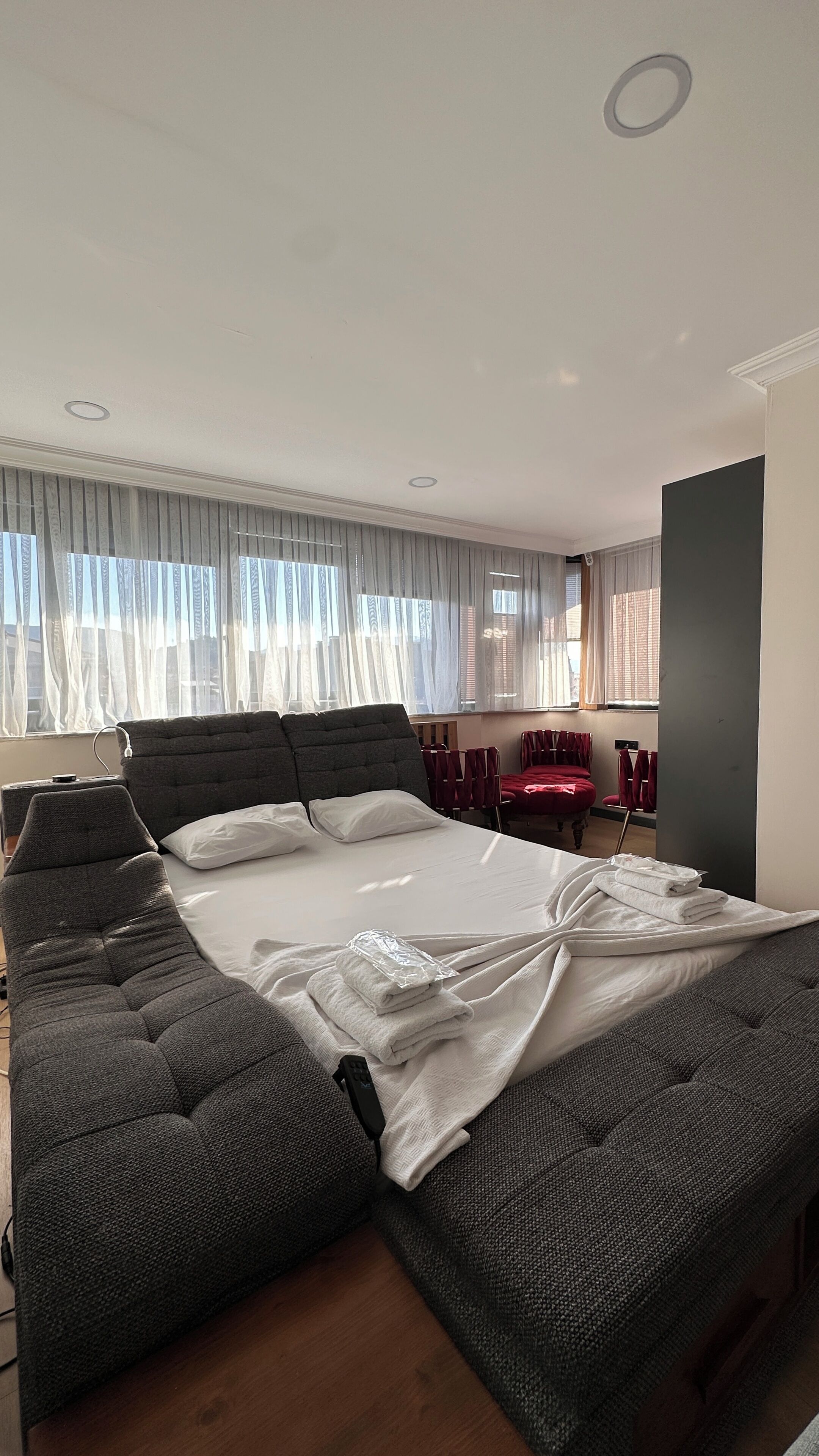 Una habitación de hotel moderna con una cama grande, una mesita de noche, una lámpara y vistas a la ciudad a través de cortinas translúcidas.