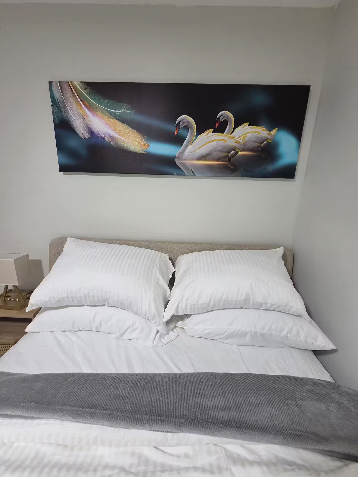 A bedroom with a bed, pillows, and a wall-mounted painting of swans.
