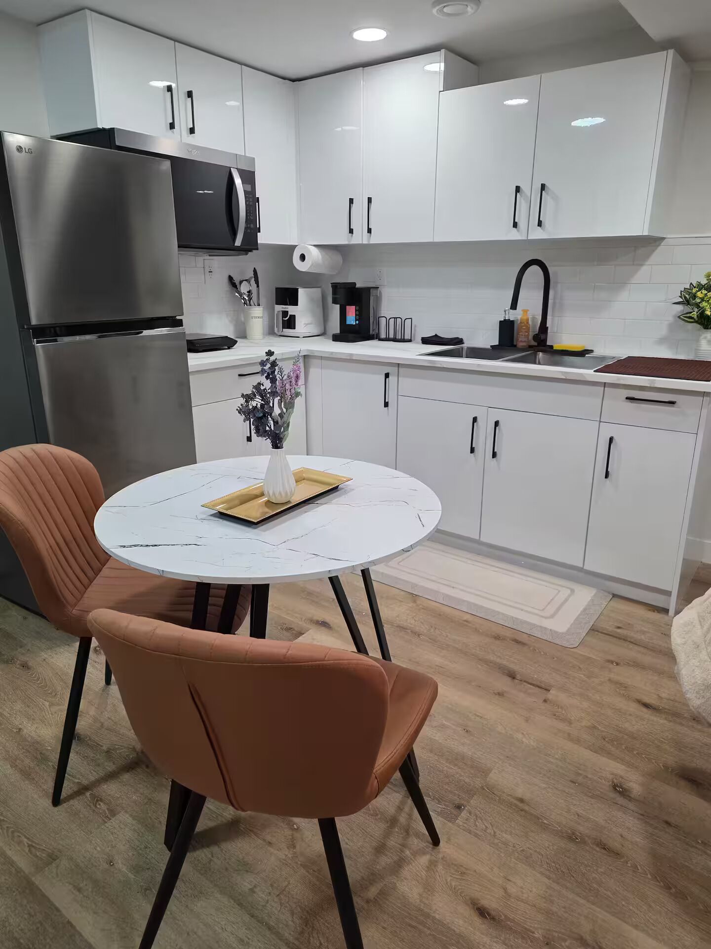 Une cuisine moderne avec des meubles blancs, un réfrigérateur en acier inoxydable, une machine à café et un coin repas comprenant une table ronde et deux chaises.