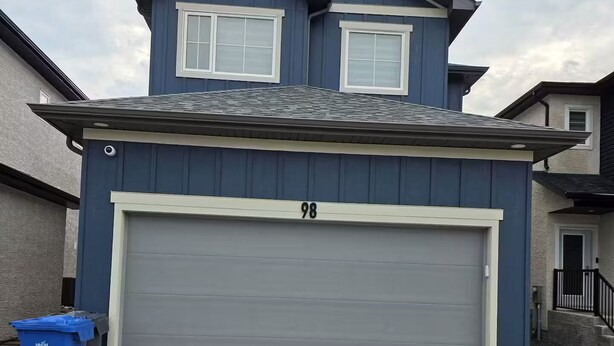 Une maison à deux étages avec un garage portant le numéro 98, une allée et une poubelle bleue pour le recyclage.
