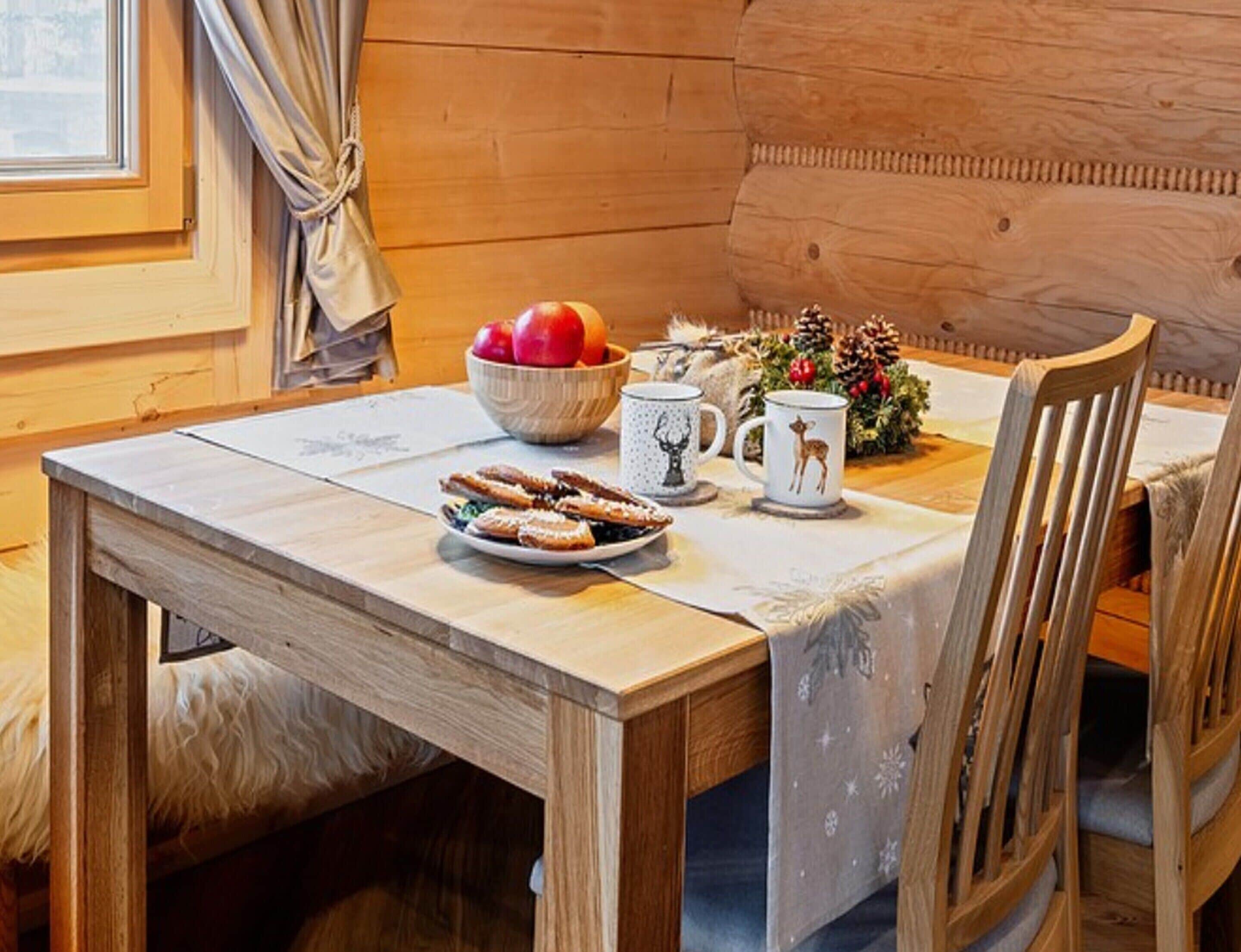 A wooden dining table with a white tablecloth, a bowl of fruit, two mugs, and a plate of pastries.