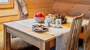 A wooden dining table with a white tablecloth, a bowl of fruit, two mugs, and a plate of pastries.