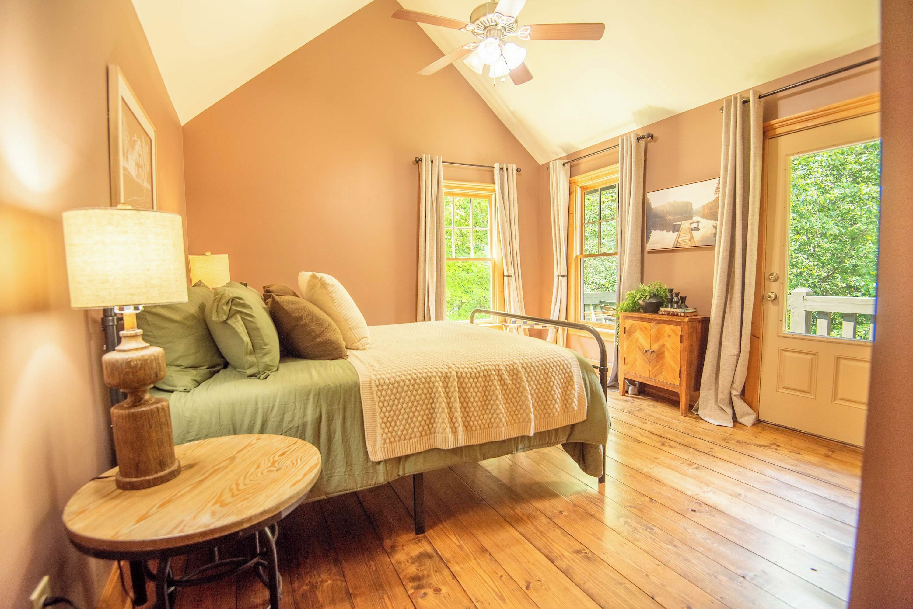 A bedroom with a bed, bedside table, lamp, and a view of the outdoors through a window.