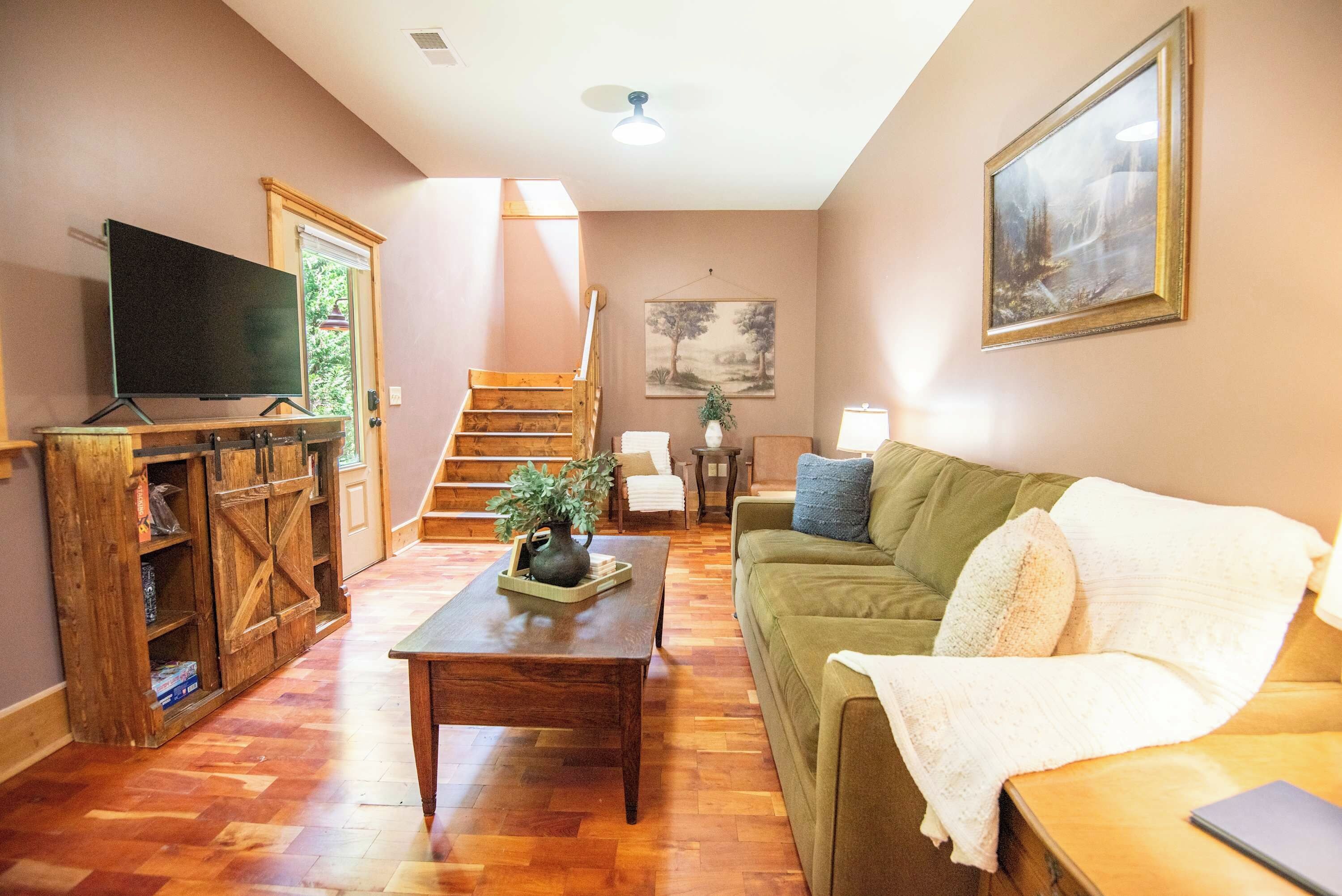 A living room with a wooden coffee table, a green sofa, a television on a stand, a staircase, and a painting on the wall.