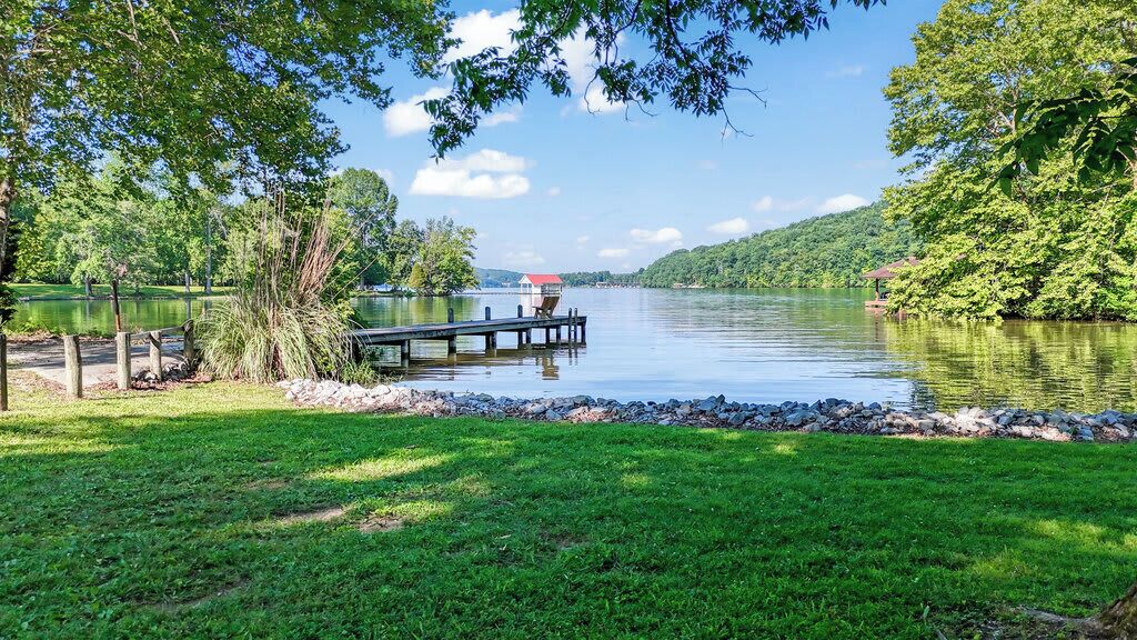 A lakeside view with a dock extending into the water, surrounded by lush greenery and a clear sky.