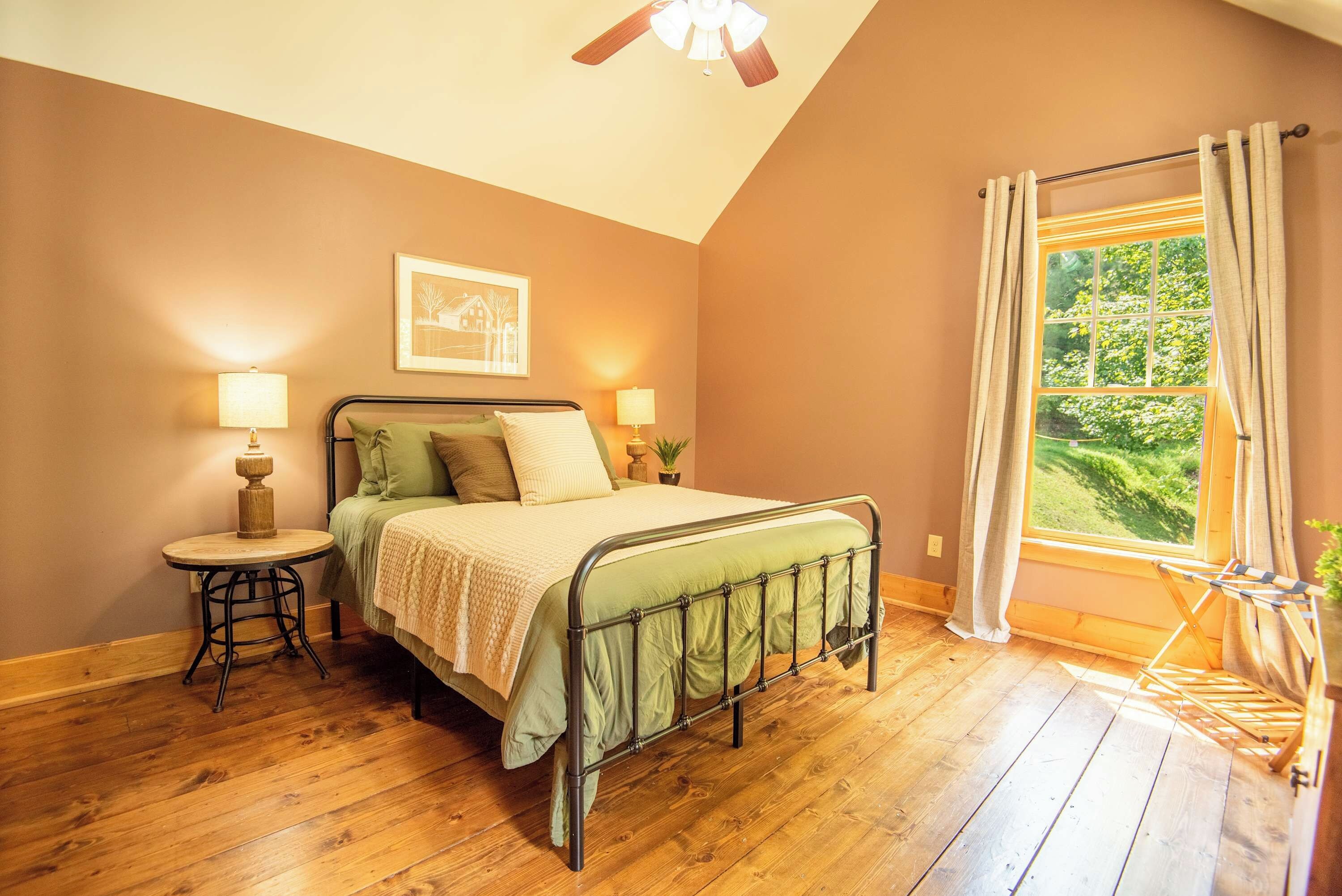Une chambre à coucher avec un lit, des lampes de chevet, un plancher en bois, un ventilateur de plafond, une fenêtre avec des rideaux et un tableau encadré au mur.
