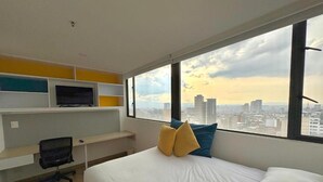 A hotel room with a bed, a TV on a shelf, and a city view. - Hotel Happa Hub (Bogota)