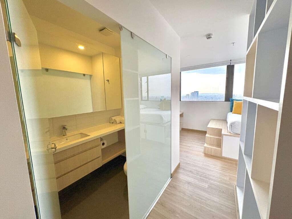 A modern hotel room with a glass partition, a large window with a city view, a wooden floor, and a white bed.
