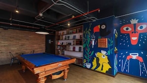 A pool table in a room with a wooden floor, a brick wall, and a bookshelf with various items. - Hotel Happa Hub (Bogota)