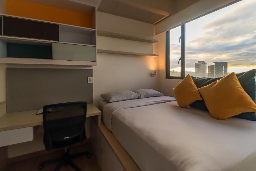 A modern hotel room with a bed, a desk, and a chair. There is a window providing a view of the city skyline.