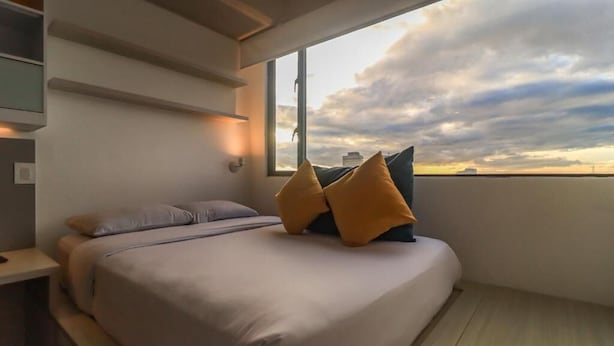 A bedroom with a bed, pillows, a nightstand, and a window with a view of the sky. - Hotel Happa Hub (Bogota)
