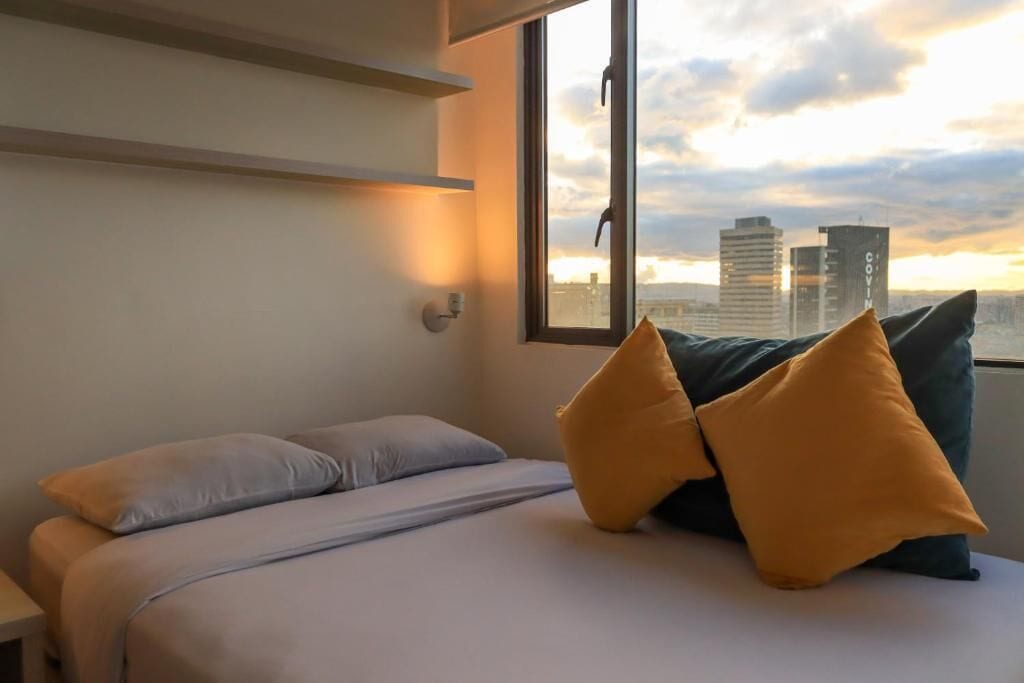 Et hotelværelse med seng, puder, et natbord og et vindue med udsigt over bybilledet.