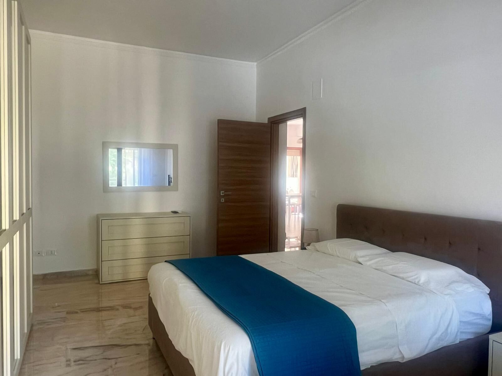 Ein Schlafzimmer mit einem Bett, einer Kommode, einem Spiegel und einer Tür zu einem weiteren Raum.