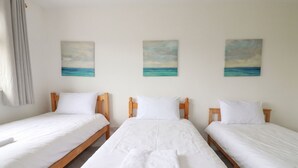 A hotel room with three single beds, each with white bedding and wooden headboards. There are three paintings of the ocean on the wall. - Cloudbreak (Wadebridge)