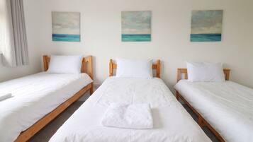 A hotel room with three single beds, each with white bedding and wooden headboards. There are three paintings of the ocean on the wall.