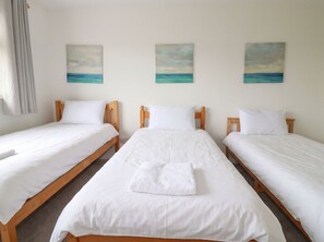 Ein Hotelzimmer mit drei Einzelbetten, jedes mit weißer Bettwäsche und Holz-Kopfteilen. An der Wand hängen drei Bilder vom Meer.