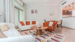 Ein modernes Wohnzimmer mit einem weißen Sofa, einem orangefarbenen Sessel, einem runden Couchtisch und einem farbenfrohen Teppich.
