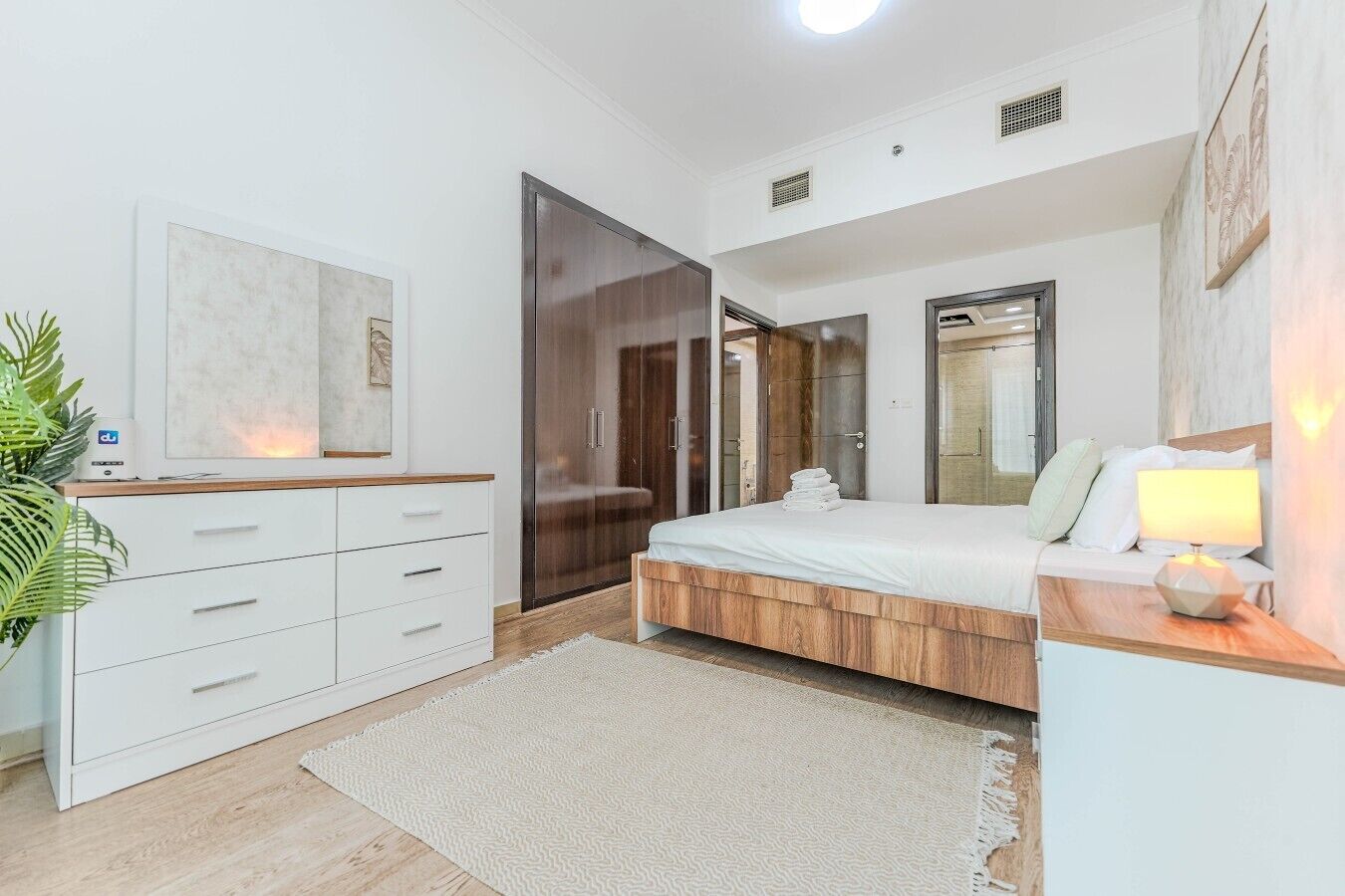 A modern bedroom with a wooden bed frame, a white bedside table with a lamp, a large wardrobe, and a wall-mounted TV.