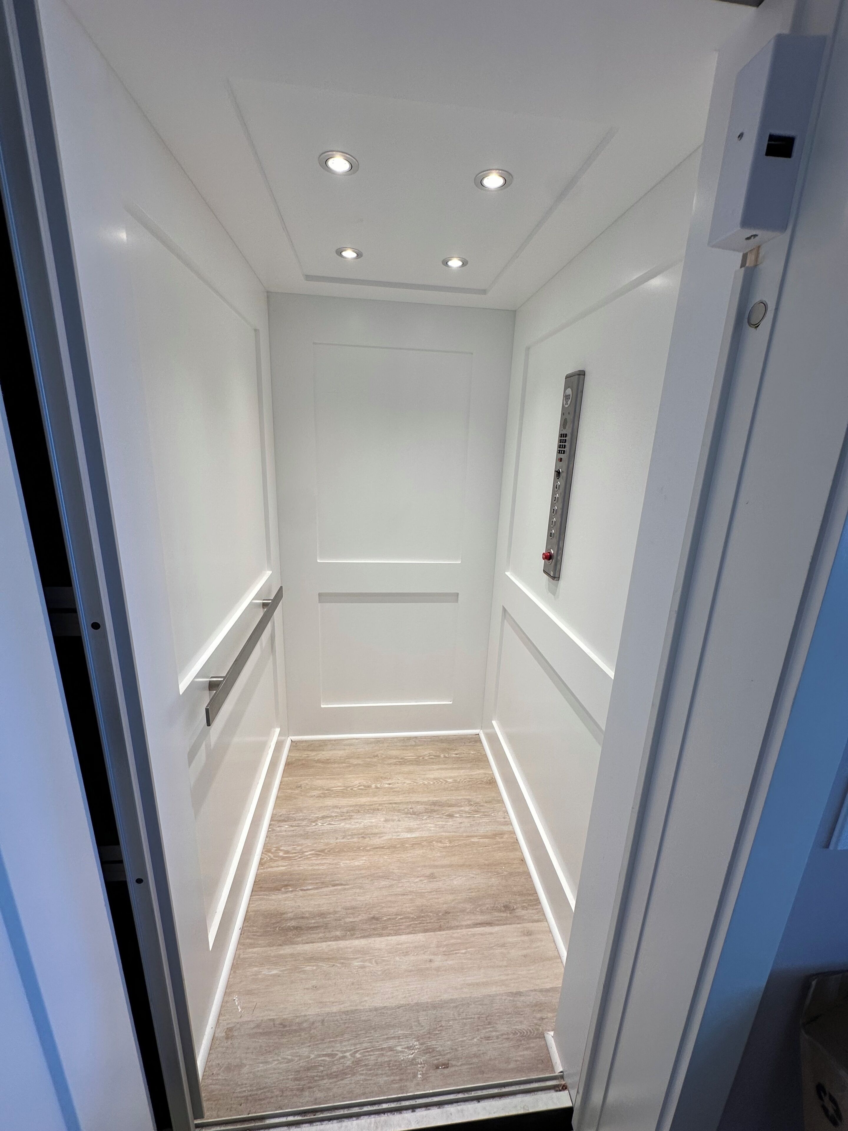 Un ascenseur étroit aux murs blancs, avec un plancher en bois et un panneau de commande situé à droite.