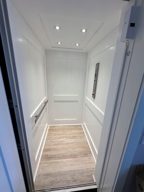A narrow elevator with white walls, a wooden floor, and a control panel on the right.