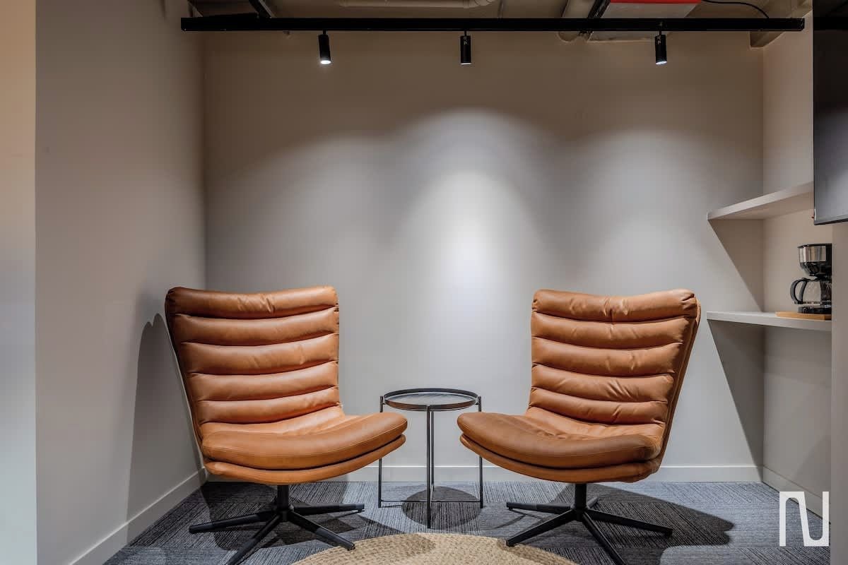 A modern room with two leather chairs and a small round table, a coffee machine on a shelf, and a visible ceiling with lighting fixtures.