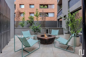 Um terraço no telhado com duas cadeiras de metal verde, uma lareira central e plantas em vasos.