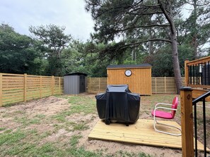 Ein Garten mit einer Holzterrasse, einem überdachten Grill, einem roten Stuhl und einem Geräteschuppen.