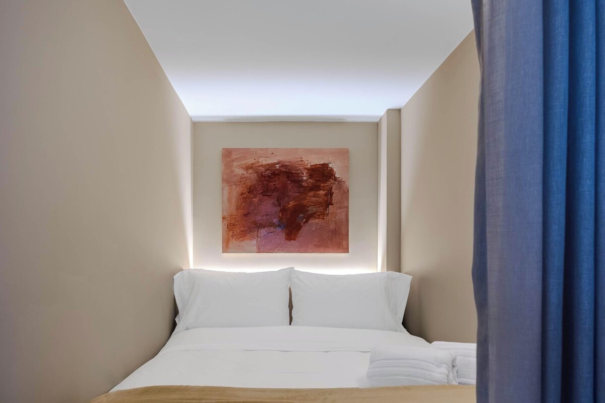 A hotel room with a bed, a wall-mounted abstract painting, and a window with blue curtains.