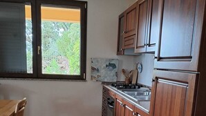 A kitchen with wooden cabinets, a tiled floor, a window with a view of greenery, and a dining area with a table and chairs. - Casa Rurale - S'Olivare di Filieri (Dorgali)
