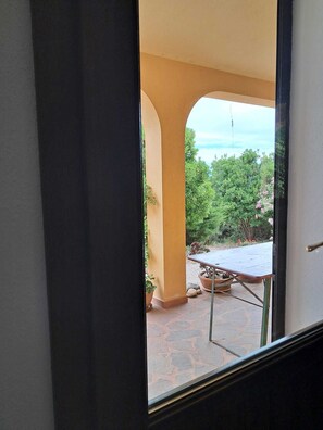 Blick durch eine Tür auf eine Außenpatio-Terrasse mit Tisch und Stühlen, umgeben von Grünpflanzen und blauem Himmel.