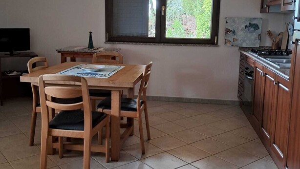 A dining area with a wooden table and chairs, a window with a view of greenery, and a kitchen with wooden cabinets. - Casa Rurale - S'Olivare di Filieri (Dorgali)