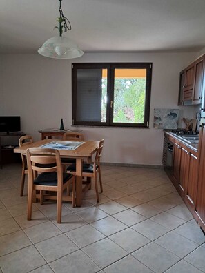 A dining area with a wooden table and chairs, a window with a view of greenery, and a kitchen with wooden cabinets. - Casa Rurale - S'Olivare di Filieri (Dorgali)