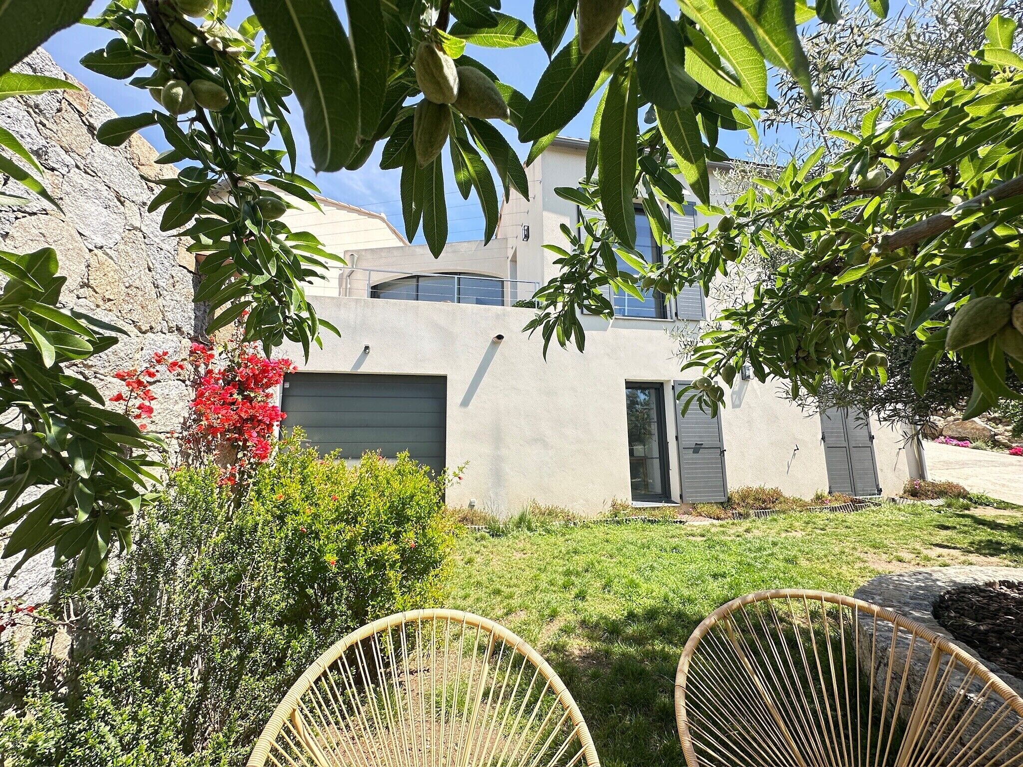 Una casa moderna con un muro de piedra, un patio cubierto de césped y dos sillas de mimbre.