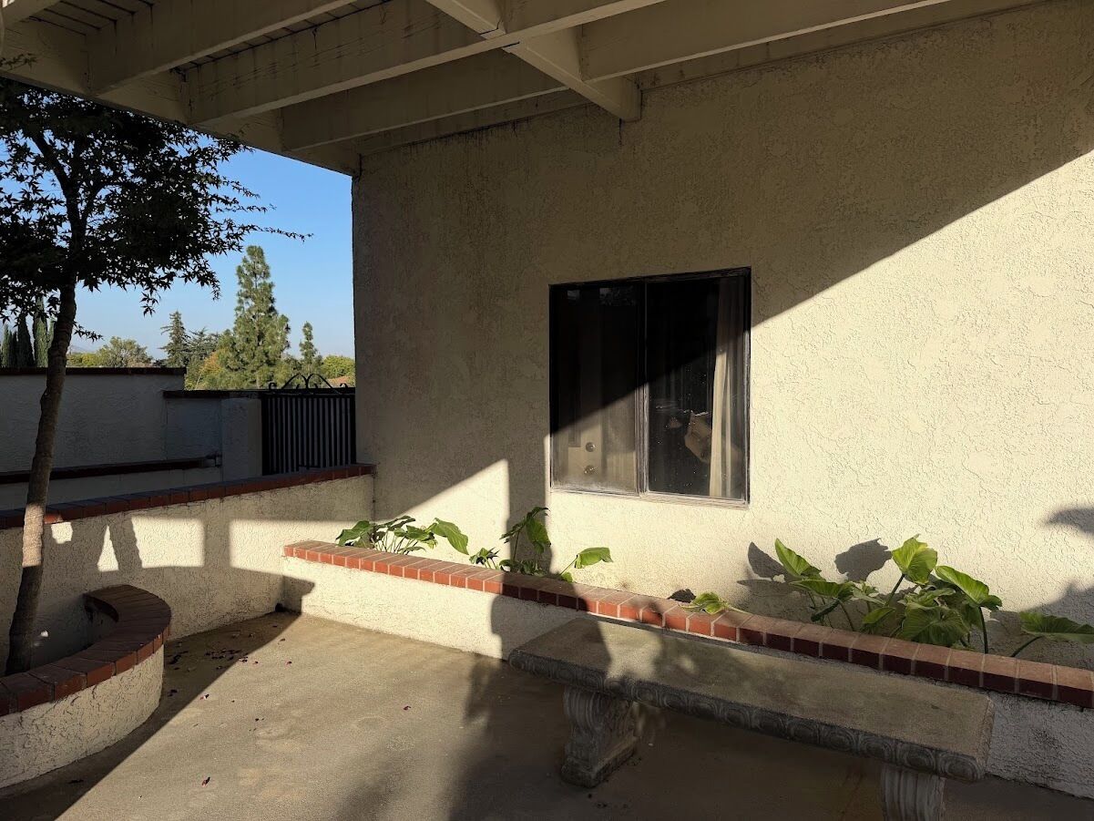 A covered outdoor area with a concrete floor, a brick planter with green plants, and a window with a dark blind.